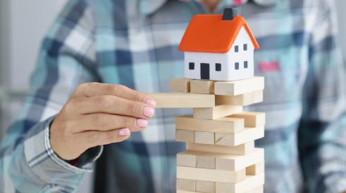 A man is taking a block out of a jenga tower with a mini house on top