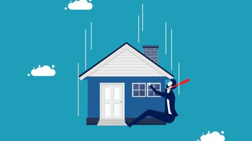 Businessman in suit holding onto falling house with blue background