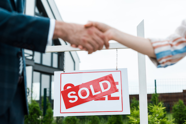 Broker and buyer shaking hands with a home sold sign behind them