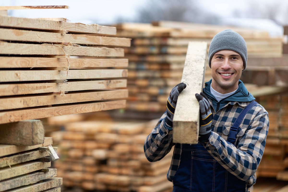 A man is holding a board of lumber with stacks of lumber behind him.
