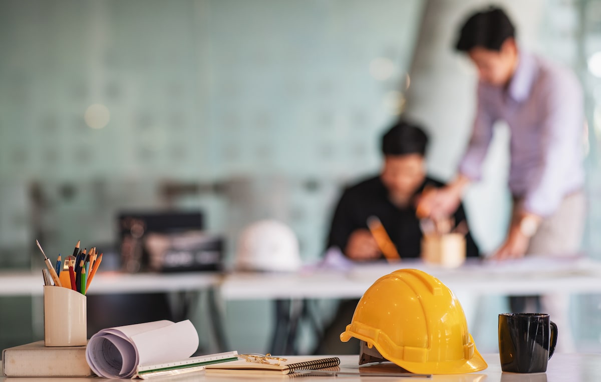 Hard hat on desk with pens and floor plans with students in background