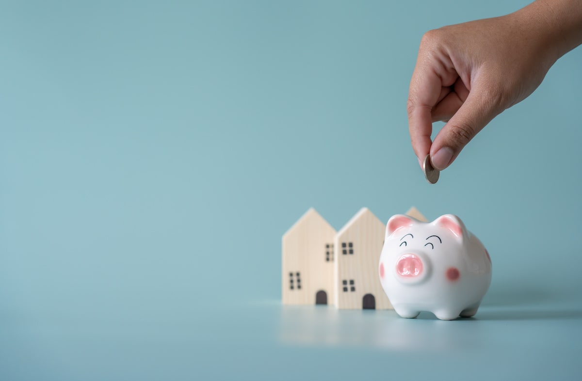 Person putting coin in piggy bank next to small wooden houses