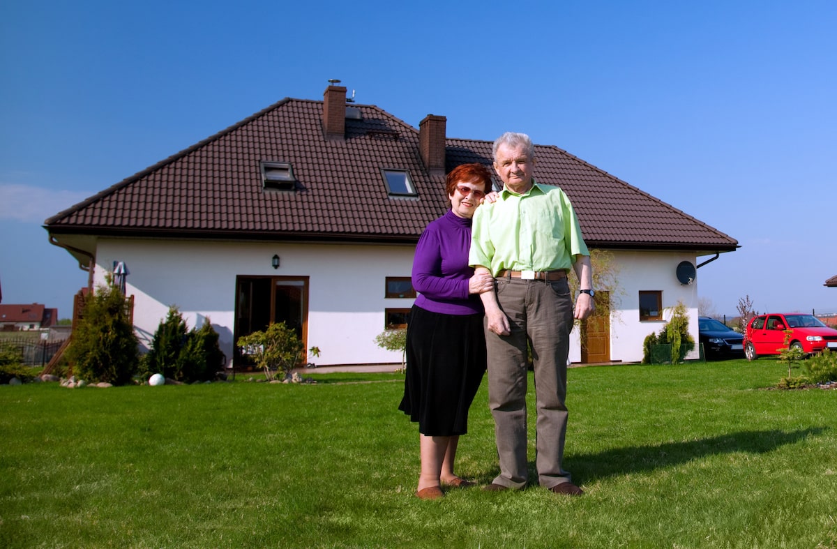 Old couple standing on lawn outside of white residential home