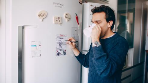Young man writing reminder on fridge and drinking coffee at home