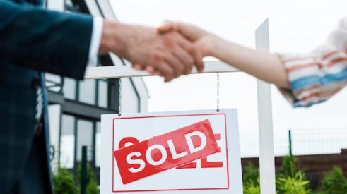 Broker and buyer shaking hands with a home sold sign behind them