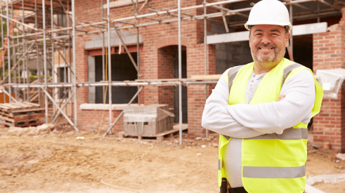 Happy home builder who enjoys his work standing in front of brick home under construction