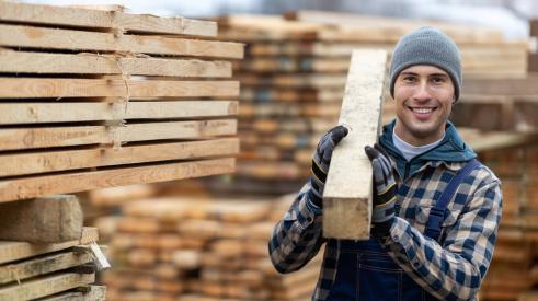 A man is holding a board of lumber with stacks of lumber behind him.