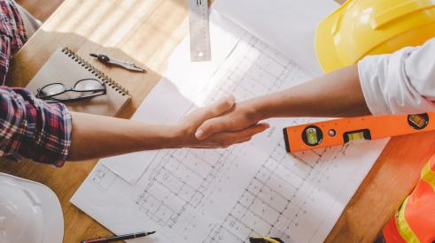 Contractor and client shaking hands above desk with paperwork on it