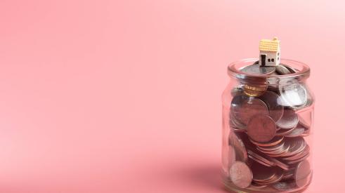 Small house model on top of jar filled with coins with pink background