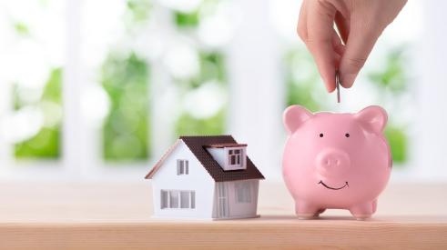 Person putting coin in piggy bank next to small house
