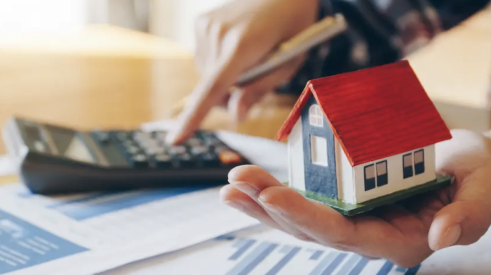 Man holding home figurine in one hand while using calculator to calculate home mortgage with the other hand 