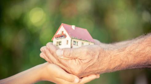 Old and young hands holding multigenerational home model against spring background