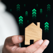 Hand holding wooden house with upward green arrows to indicate increased green building practices